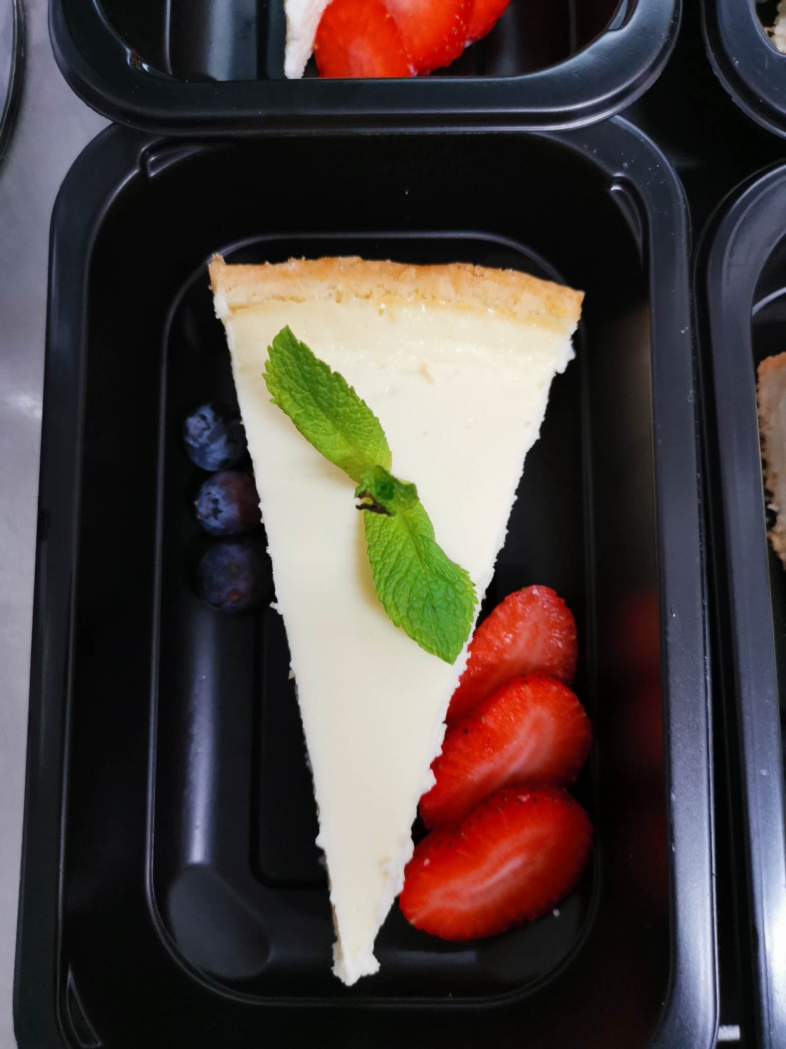 Kawałek sernika nowojorskiego z dekoracją z listka mięty, trzech borówek i trzech plastrów truskawki, umieszczony w czarnym, plastikowym pojemniku na żywność.