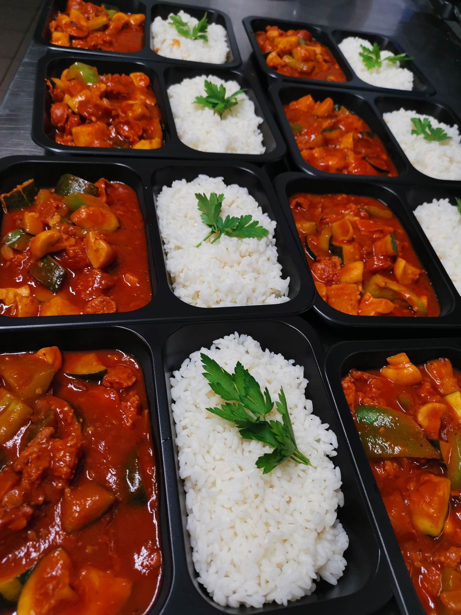 Rzędy czarnych pojemników z dwukomorowym podziałem, wypełnionych gęstym gulaszem warzywnym w czerwonym sosie i porcją białego ryżu, udekorowanego gałązką świeżej zielonej pietruszki.