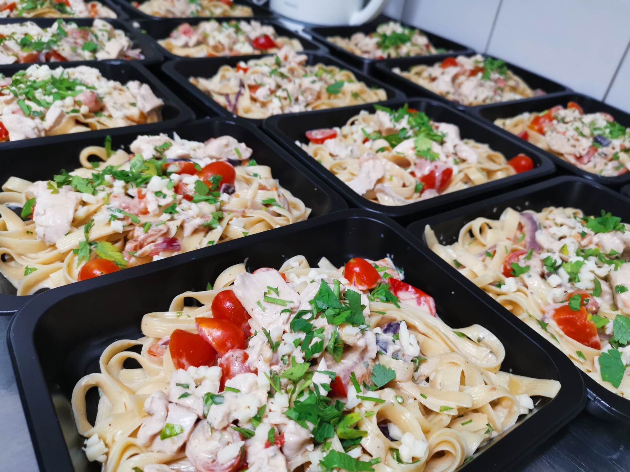 Gotowe porcje tagliatelle z kurczakiem, pomidorkami cherry i ziołami w czarnych pojemnikach cateringowych, przygotowane do dostawy w Poznaniu.