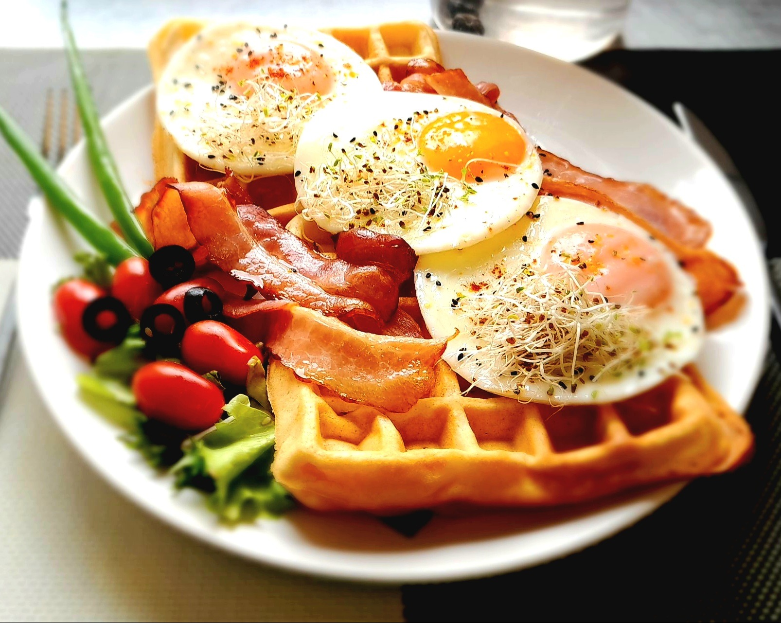 Apetycznie wyglądający posiłek: gofry z jajkami sadzonymi posypanymi kiełkami, chrupiącym bekonem, pomidorkami koktajlowymi, oliwkami i zielonym szczypiorkiem, podany na białym talerzu.