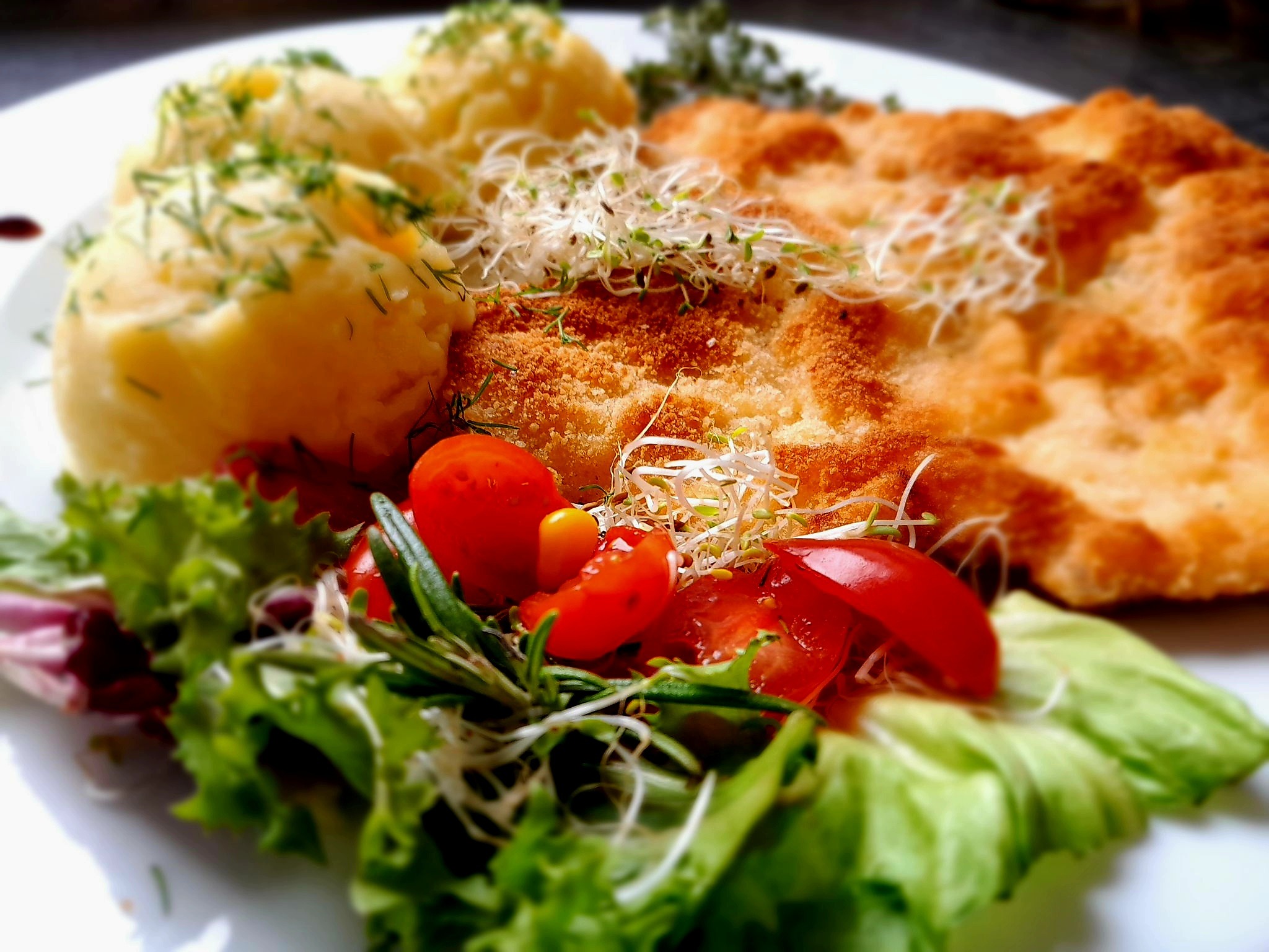 Kotlet schabowy w panierce z ziemniakami puree posypanymi koperkiem, sałatką ze świeżych pomidorów i kiełków, udekorowany gałązką rozmarynu na białym talerzu.