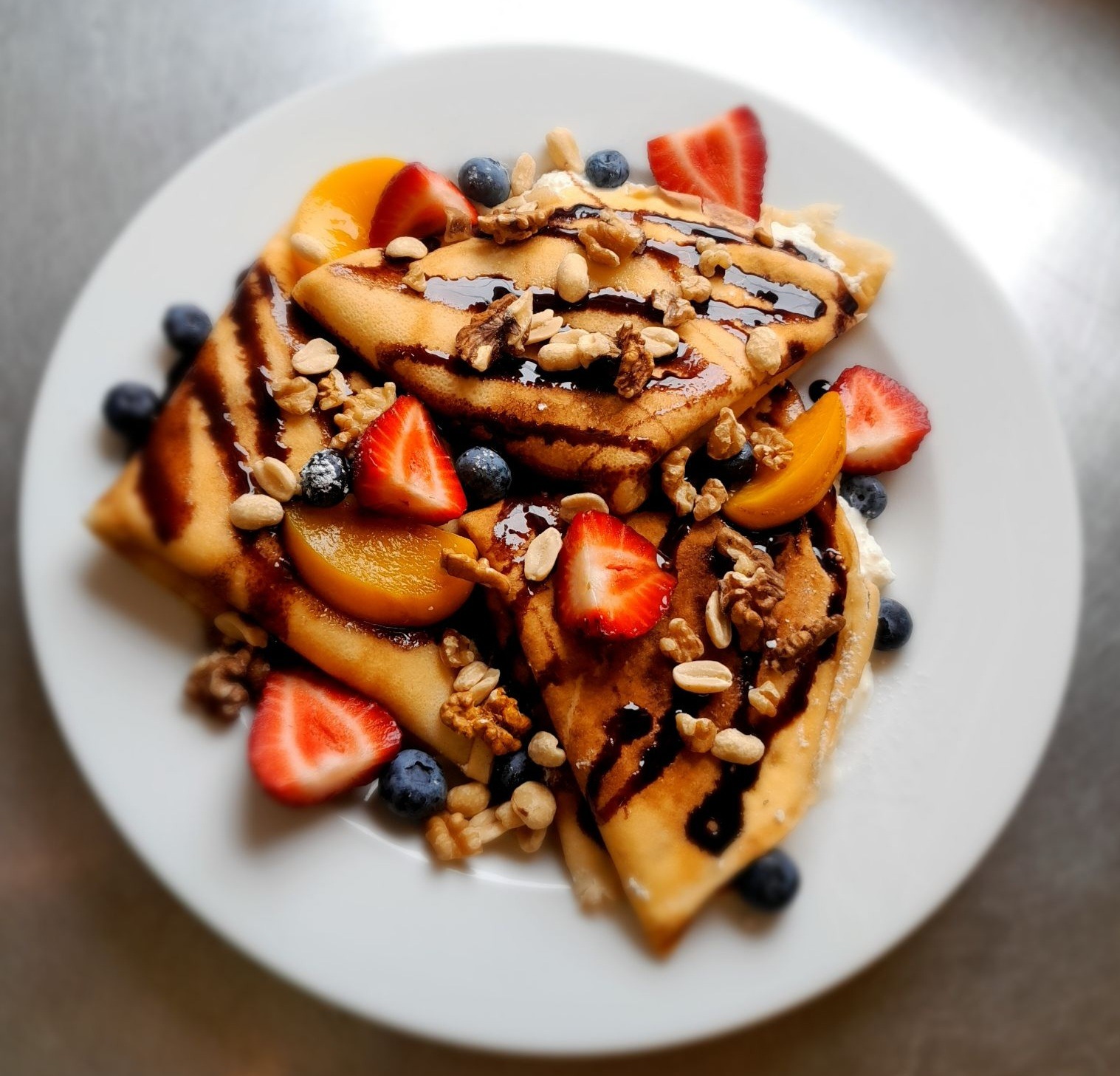 Trzy złożone naleśniki na białym talerzu, polane ciemną polewą i posypane orzechami, z dodatkiem świeżych truskawek, borówek i kawałków brzoskwini.