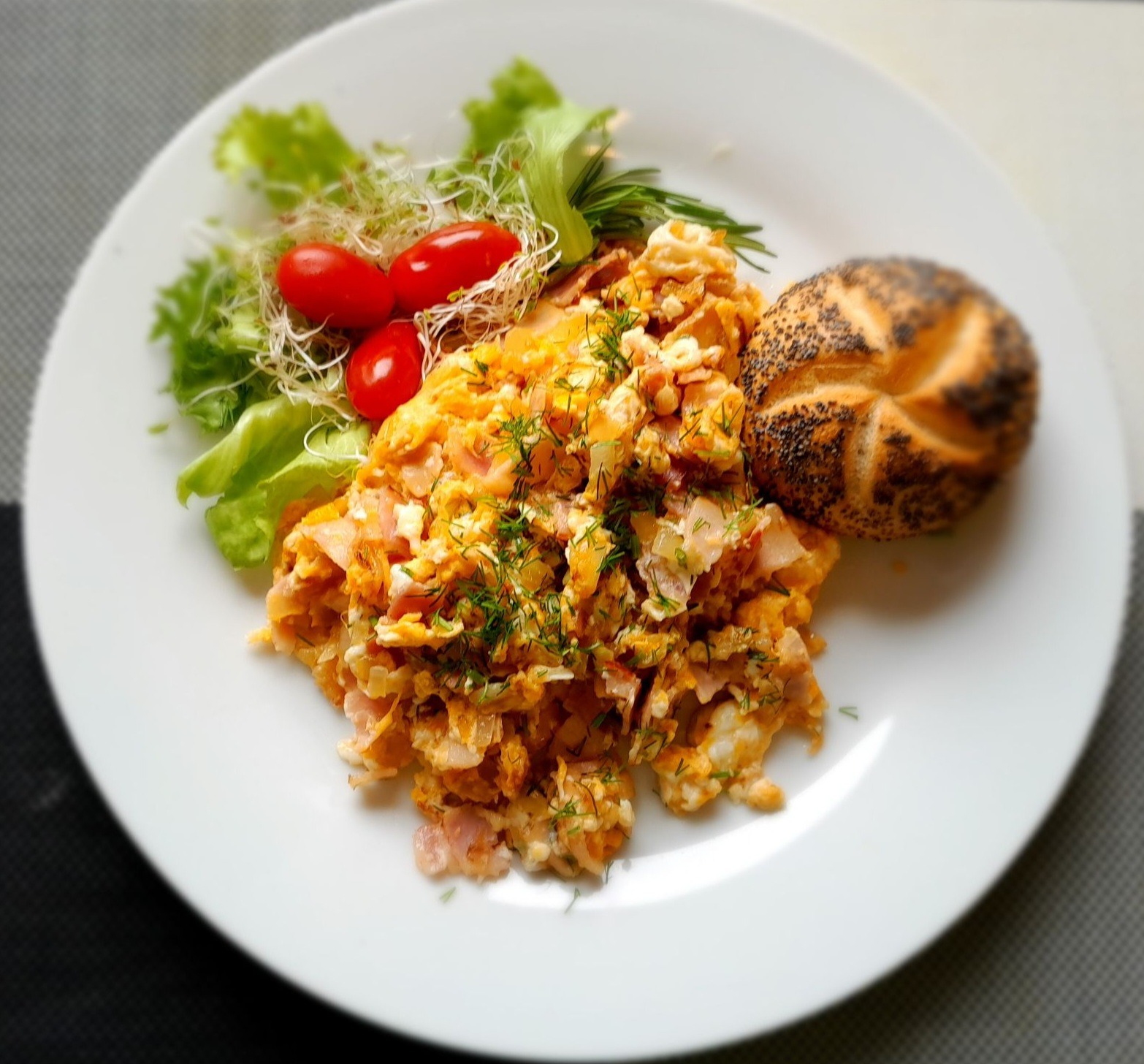 Jajecznica z szynką na białym talerzu, udekorowana sałatą, kiełkami, pomidorkami cherry i gałązką rozmarynu, podana z bułką posypaną makiem.