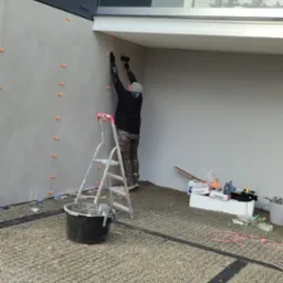 Pracownik na drabinie wygładza ścianę zewnętrzną budynku, widoczne pomarańczowe krzyżyki dystansowe, wiadro z zaprawą i narzędzia na podłożu z kruszywa.