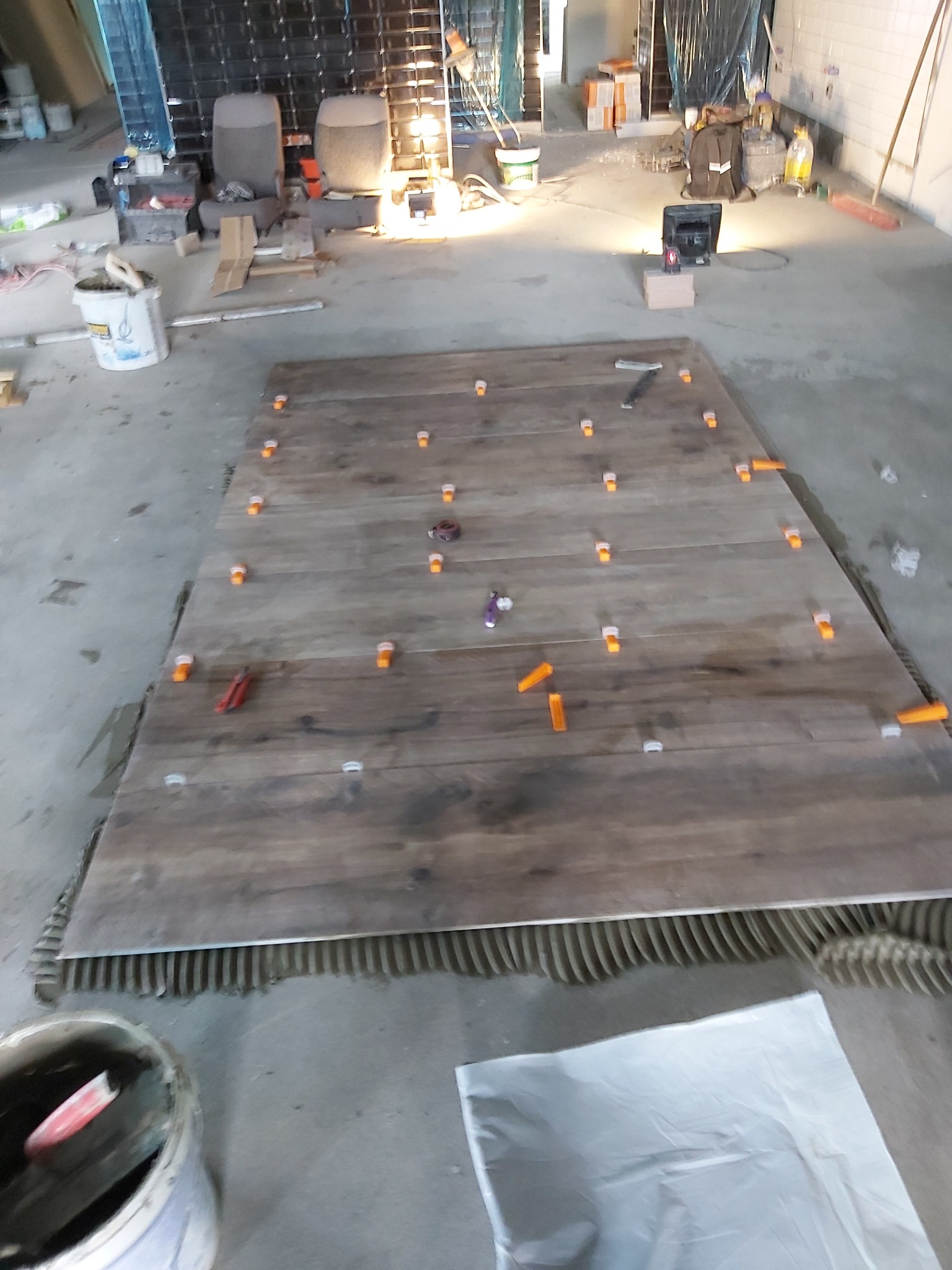 Układanie płytek imitujących drewno na podłodze, z użyciem pomarańczowych klipsów dystansowych, widoczny klej do płytek i narzędzia w tle.