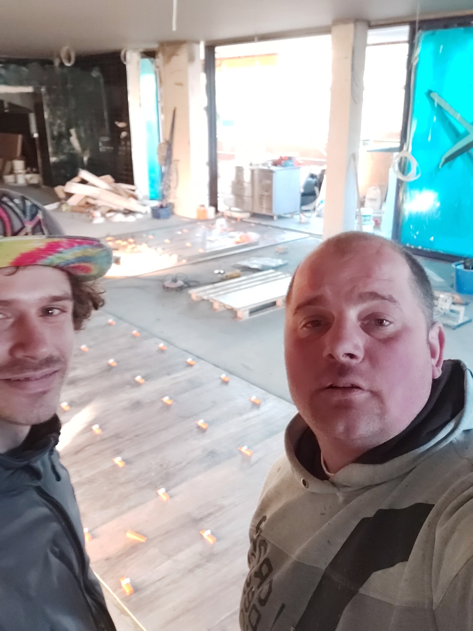 Dwóch mężczyzn pozuje na tle podłogi z płytek w trakcie układania, widoczne plastikowe krzyżyki dystansowe i elementy wykończeniowe w tle.