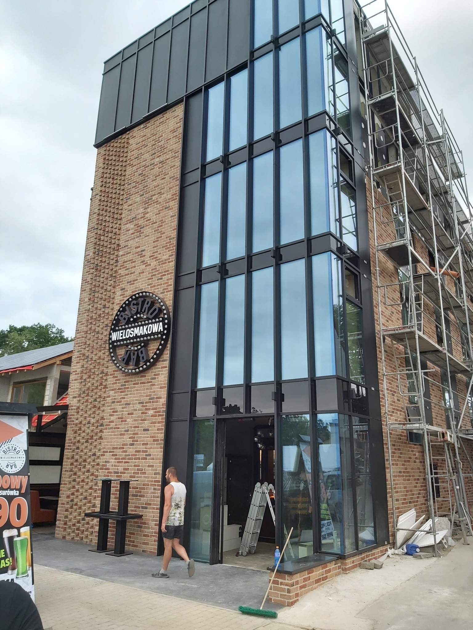Nowoczesny budynek bistro z ceglaną elewacją, dużymi oknami i metalowymi elementami, częściowo w trakcie prac wykończeniowych z widocznym rusztowaniem i narzędziami.