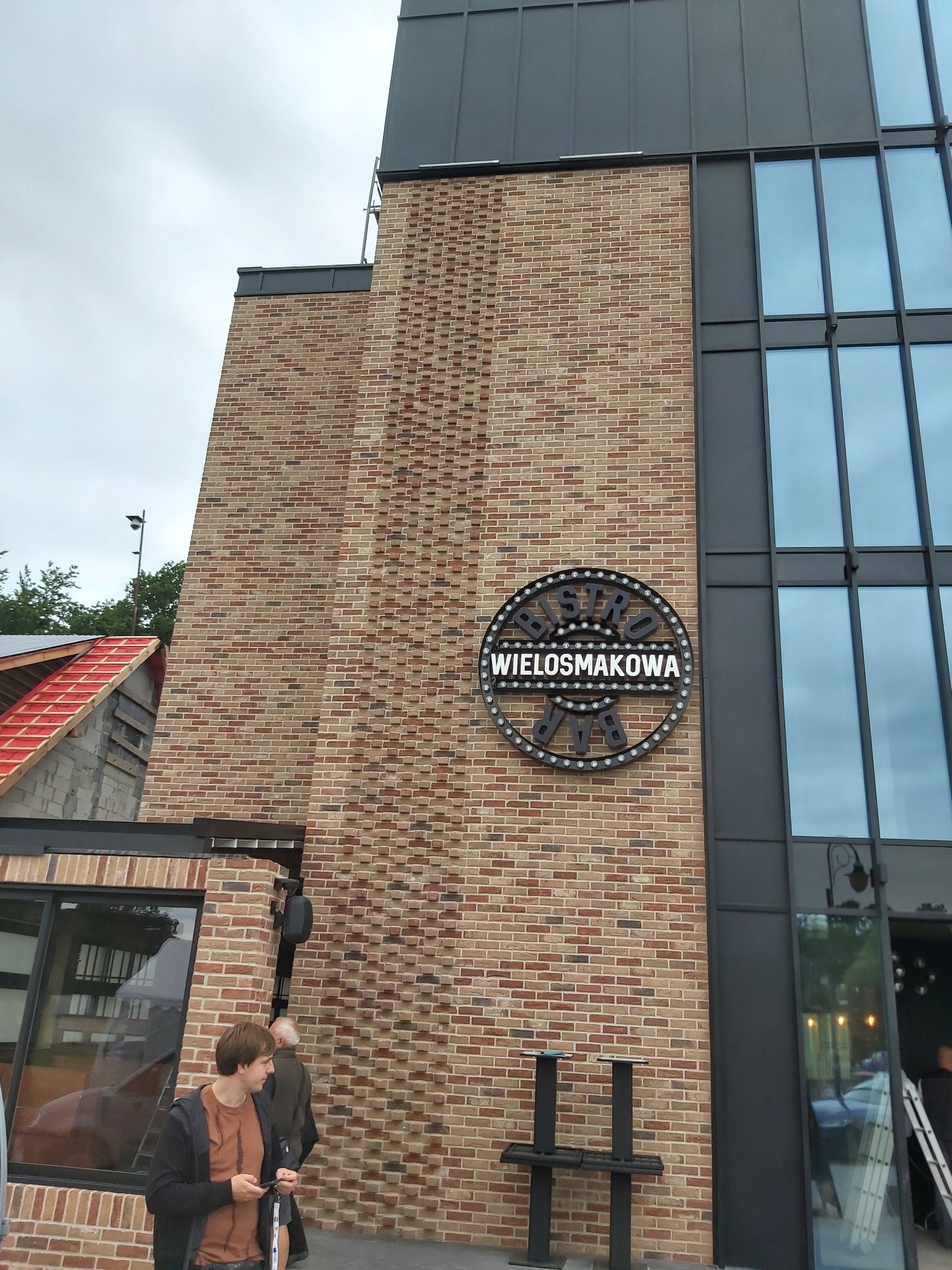 Murowana elewacja budynku z cegły klinkierowej w odcieniach brązu i beżu, z dużymi oknami w czarnych ramach i okrągłym szyldem z nazwą 'Bistro Wielosmakowa Bar'.