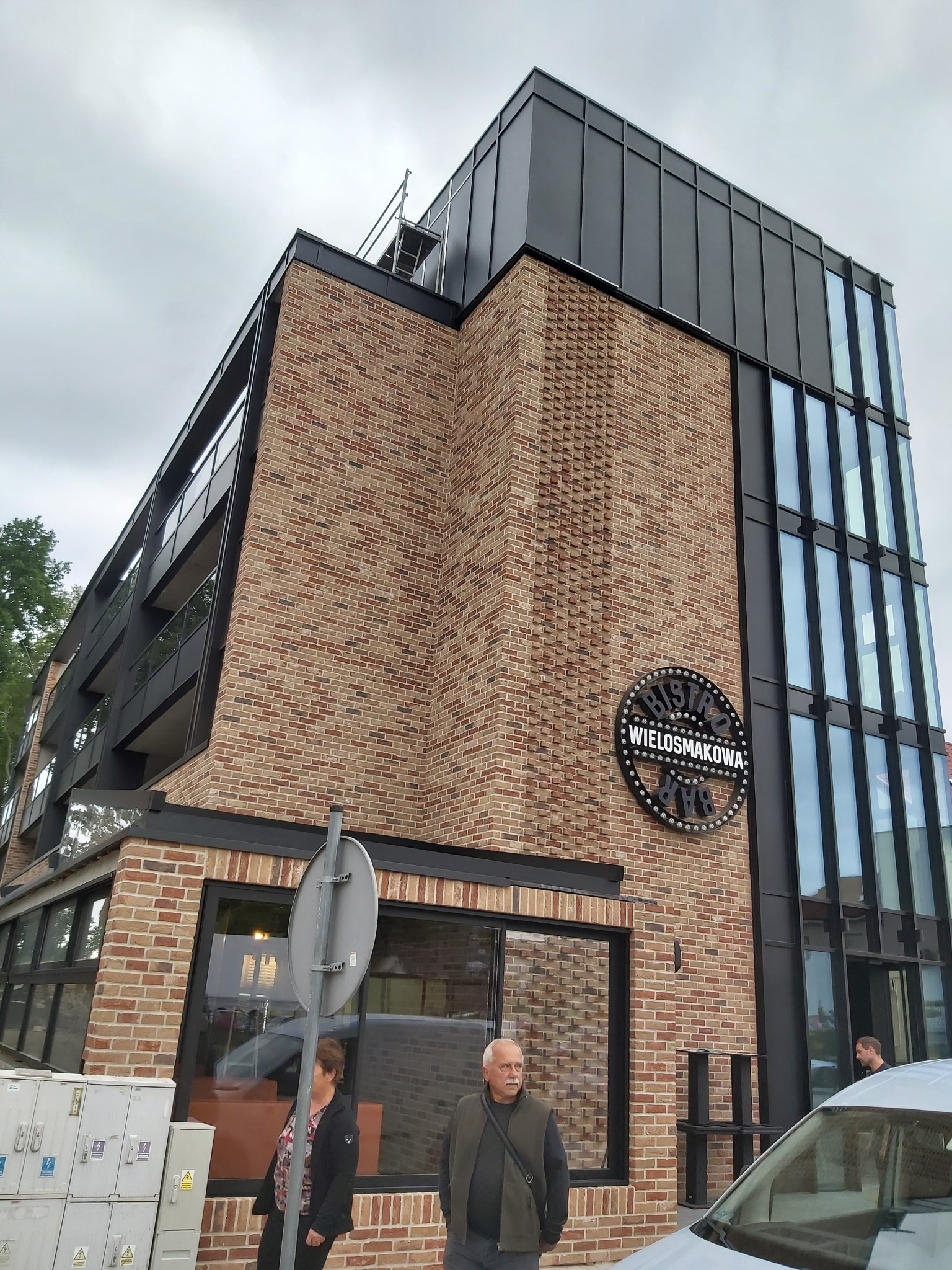 Nowoczesny budynek z cegły klinkierowej z czarnymi elementami wykończeniowymi i dużymi oknami, ozdobiony okrągłym szyldem 'Bistro Wielosmakoowa Bar'.
