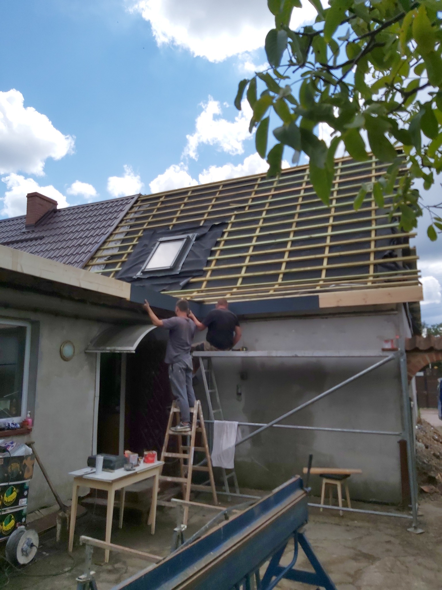 Dwóch mężczyzn na rusztowaniu montuje membranę dachową i drewniane łaty na dachu domu jednorodzinnego z brązową dachówką, widoczne narzędzia i akcesoria na stole obok drabiny.