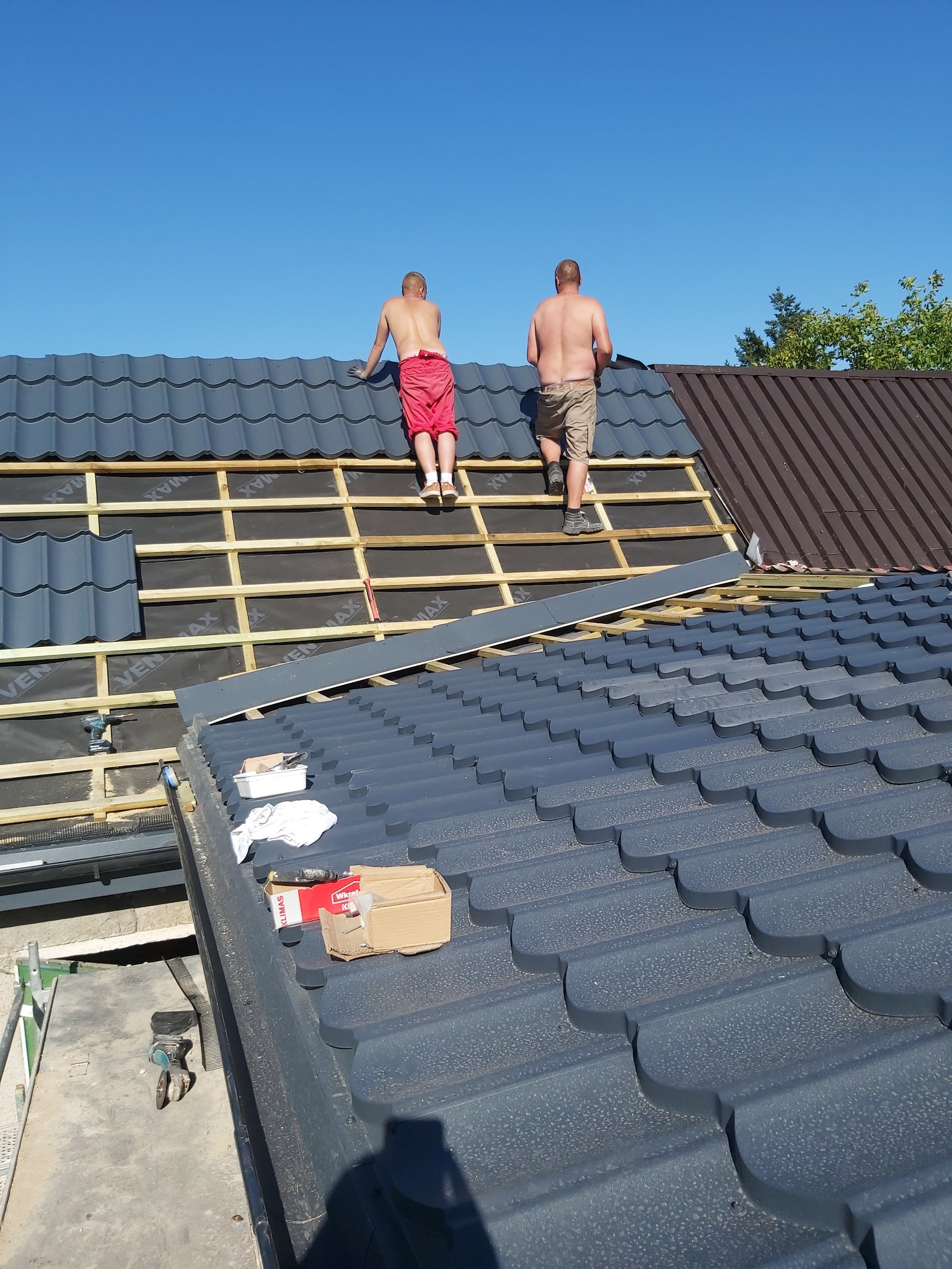 Dwóch mężczyzn bez koszulek układa ciemnoszarą dachówkę na drewnianej konstrukcji dachu, widoczna czarna membrana, fragment brązowej dachówki po prawej stronie.