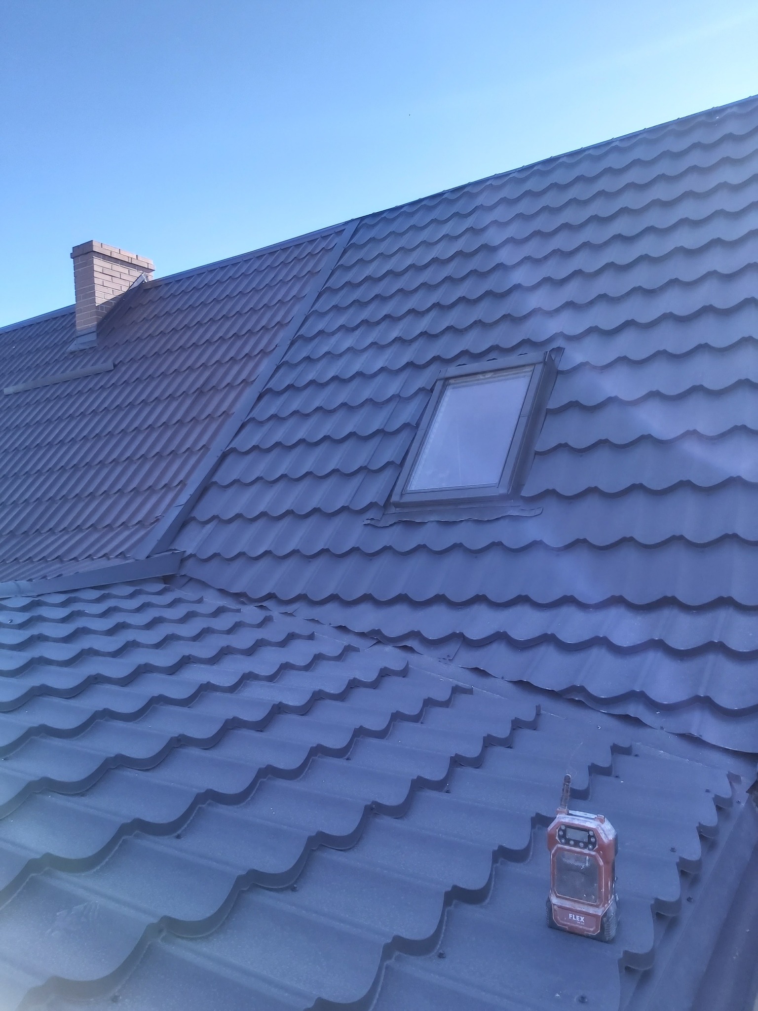 Ciemnoszara blachodachówka z oknem dachowym i ceglanym kominem w tle, na dachu stoi radio budowlane marki Flex.