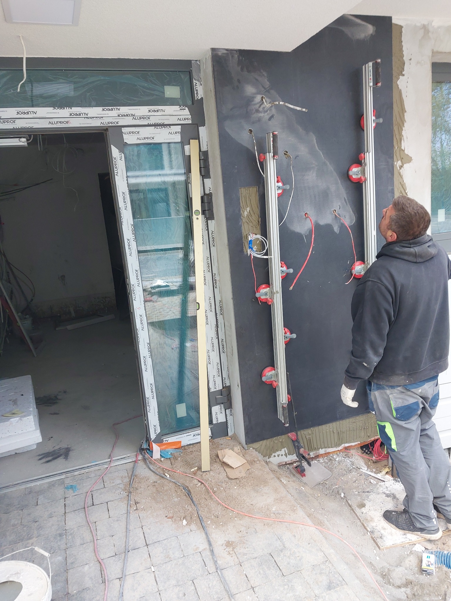 Pracownik instaluje duże okno z ramą aluminiową, używając specjalistycznych przyssawek montażowych do przenoszenia i pozycjonowania szyby, widoczne przewody elektryczne i elementy konstrukcyjne...