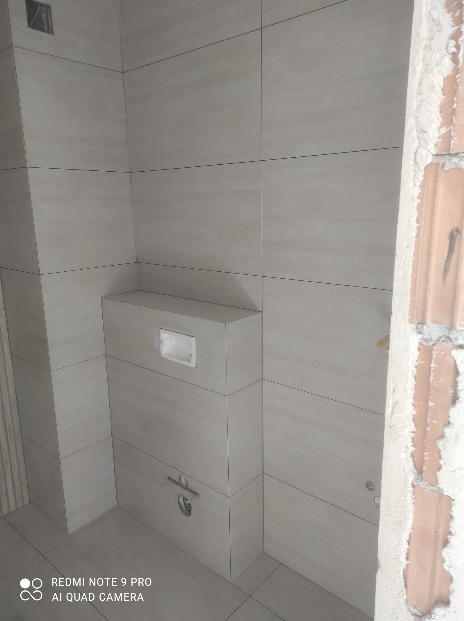 Wykończona łazienka z beżowymi płytkami, zabudowa podtynkowego WC, widoczne przyłącza, surowy mur z prawej strony kadru sugeruje etap prac wykończeniowych.