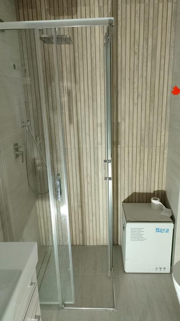 Szklana kabina prysznicowa z deszczownicą, zamontowana w łazience z płytkami imitującymi drewniane panele, obok karton z armaturą Roca.