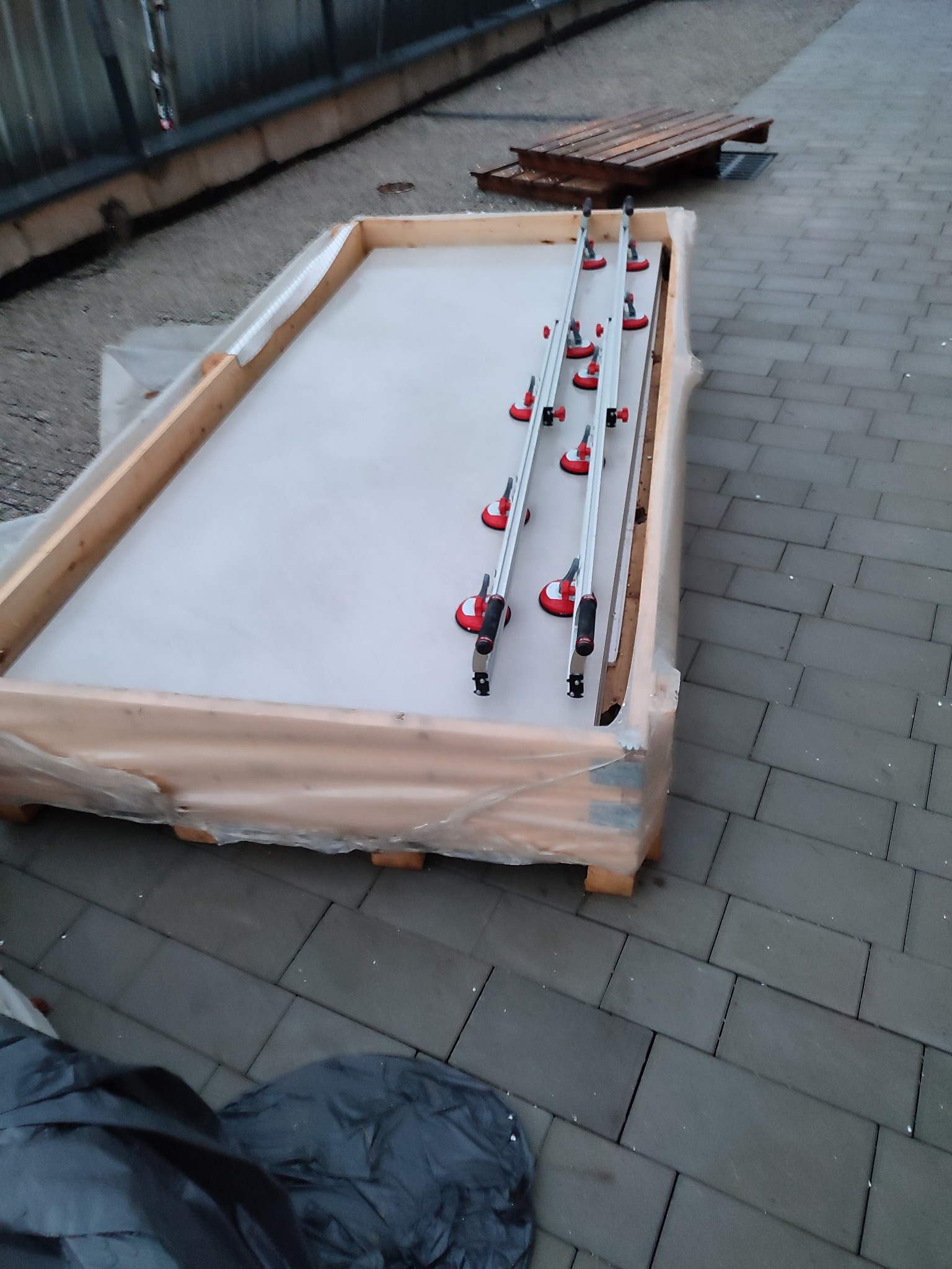 Płyta gipsowa w drewnianej ramie, zabezpieczona folią, z przyssawkami do przenoszenia, leżąca na brukowanej powierzchni.