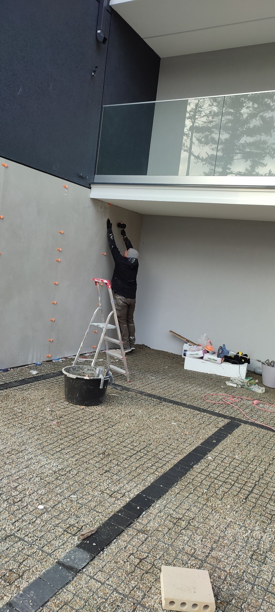 Pracownik na drabinie wygładza ścianę zewnętrzną budynku, widoczne pomarańczowe krzyżyki dystansowe, wiadro z zaprawą i narzędzia na podłożu z kruszywa.