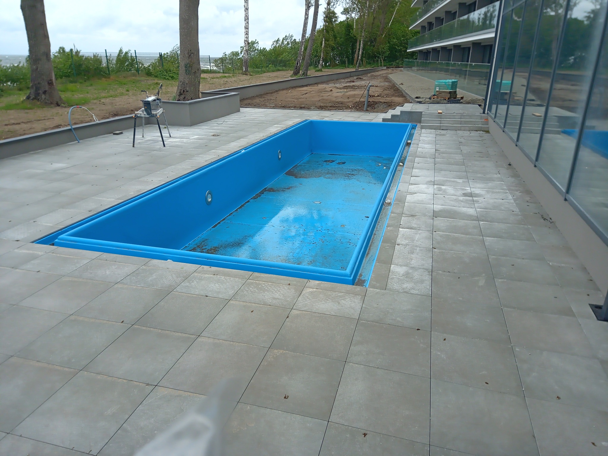 Niebieski, pusty basen w trakcie budowy lub remontu, otoczony szarą kostką brukową, widok na budynek z balkonami i drzewa w tle.