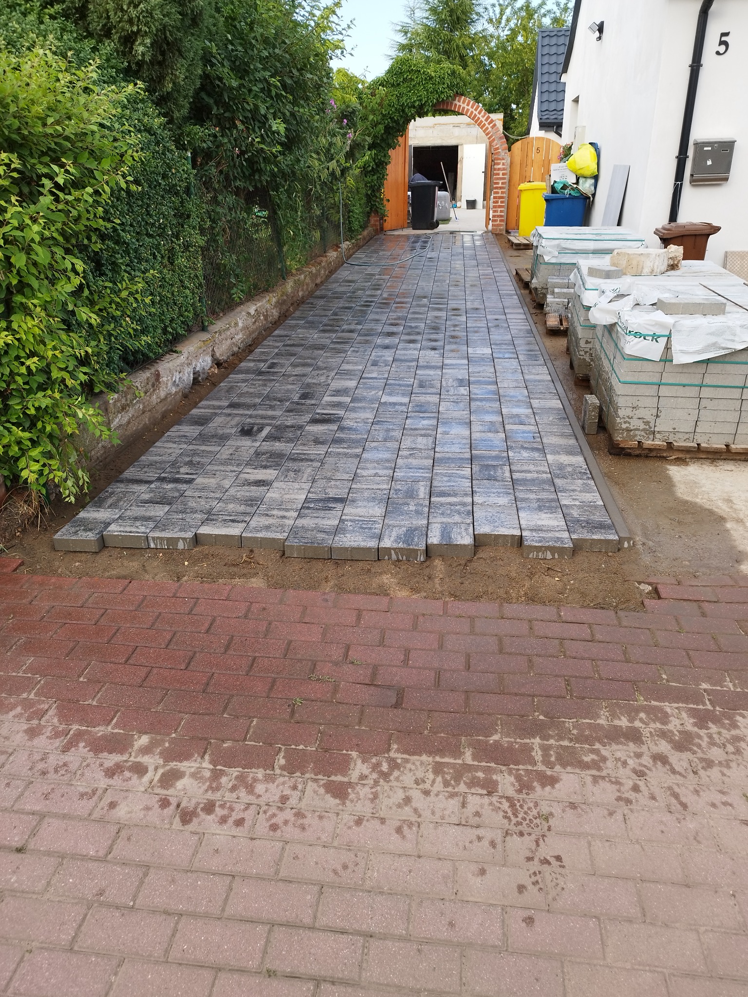 Układanie szarej kostki brukowej na podjeździe, widoczne palety z kostką, brama z ceglanym łukiem i bujna zieleń w tle.
