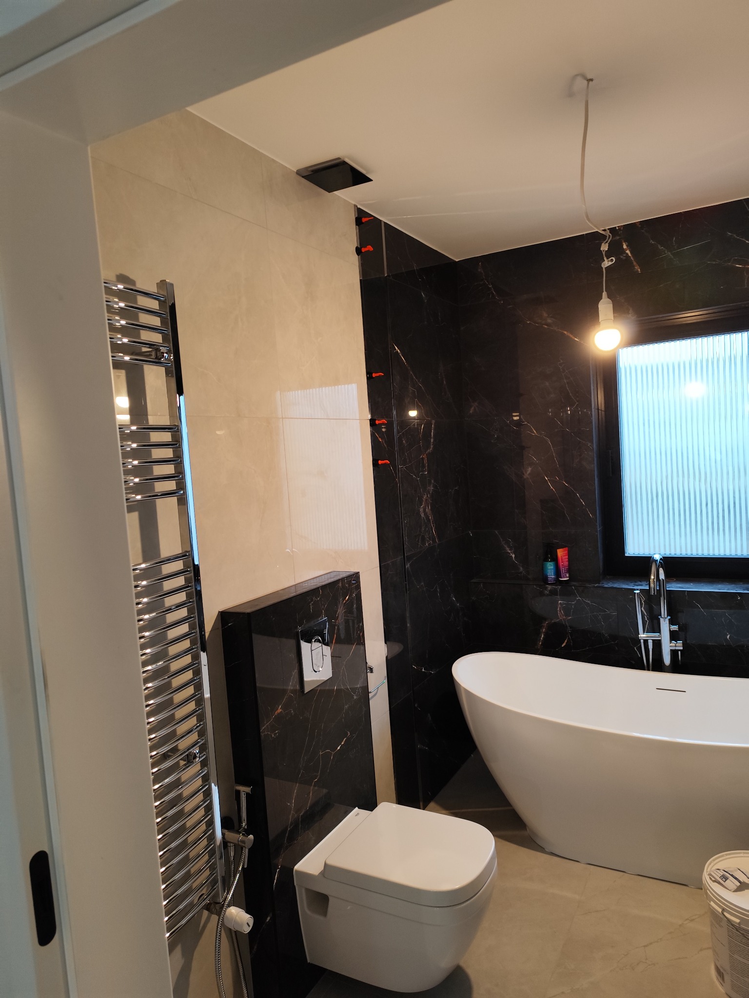 Nowoczesna łazienka z czarno-białymi marmurkowymi płytkami, owalną wanną wolnostojącą, podwieszaną toaletą i grzejnikiem drabinkowym, widoczne pomarańczowe kliny do płytek na ścianie.