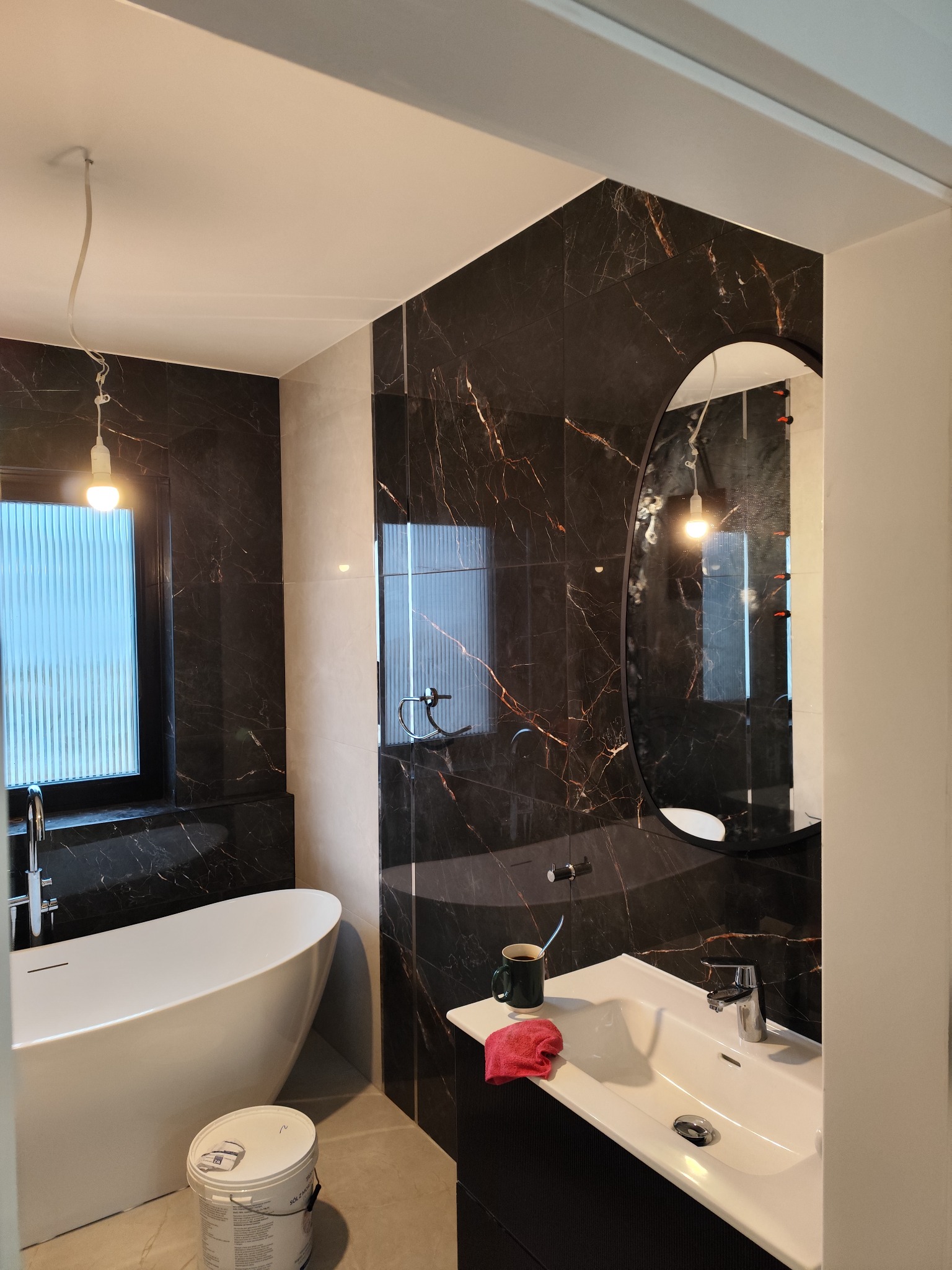 Nowoczesna łazienka z czarnymi płytkami imitującymi marmur, owalne lustro, biała wanna wolnostojąca, umywalka z szafką, kubek z szczoteczką do zębów i wiadro z farbą na podłodze.