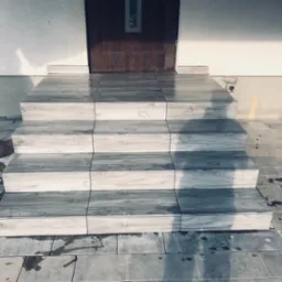 Schody wejściowe wyłożone szarymi płytkami imitującymi drewno, prowadzące do ciemnych drzwi z małym okienkiem, oświetlone naturalnym światłem.