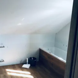 Biała wanna z hydromasażem zabudowana panelami imitującymi drewno, białe płytki na ścianach pod skosem, podłoga wyłożona panelami imitującymi drewno, widoczne narzędzia i środki czystości w wiadrze...