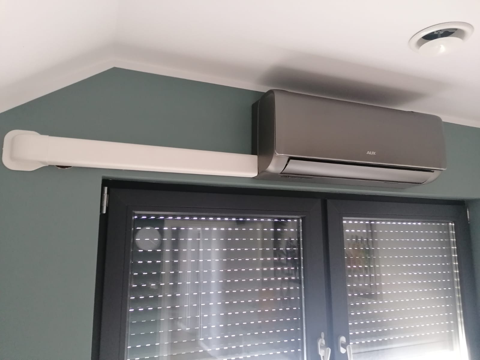 Szary klimatyzator marki AUX zamontowany nad oknem z roletami zewnętrznymi, z widoczną białą maskownicą instalacji na ścianie w kolorze morskim.