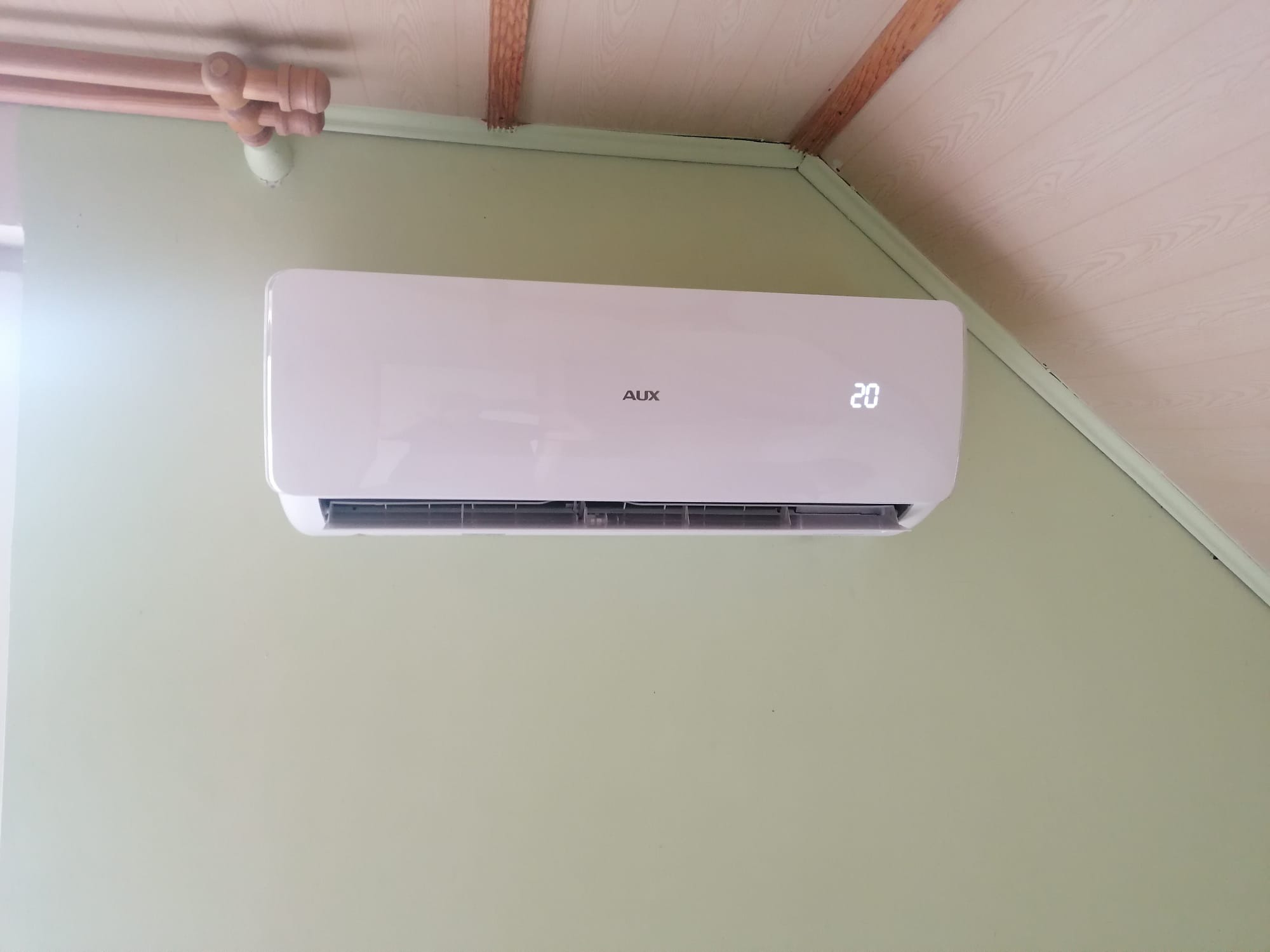 Biała jednostka klimatyzacji AUX zamontowana na jasnozielonej ścianie w pomieszczeniu z drewnianym wykończeniem sufitu, wyświetlająca temperaturę 20 stopni.