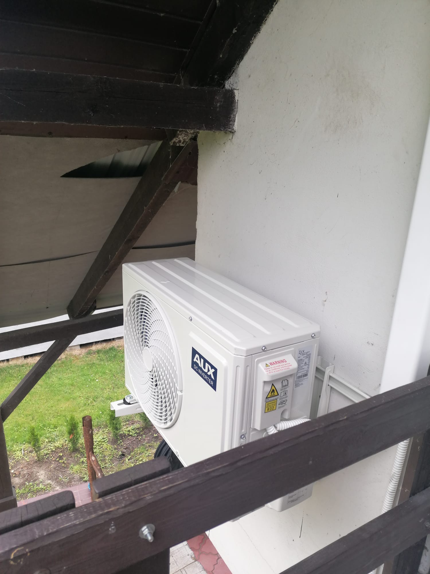 Zewnętrzna jednostka klimatyzacji AUX DC Inverter zamontowana na białej ścianie budynku, widoczna zza drewnianej balustrady z widokiem na trawnik.