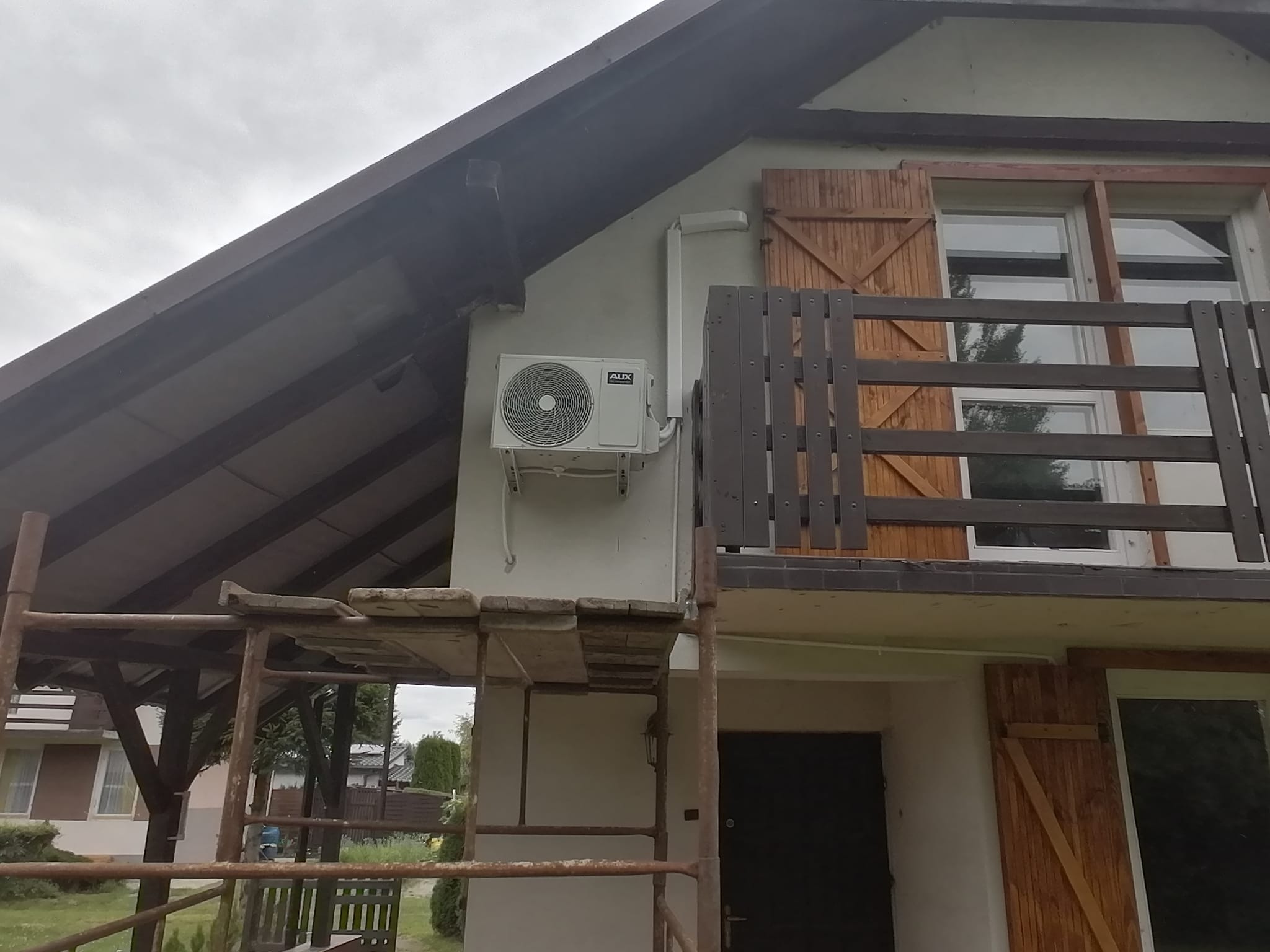 Biała jednostka zewnętrzna klimatyzacji marki AUX zamontowana na ścianie budynku z drewnianymi okiennicami, widoczne rusztowanie.