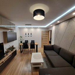 NEW DESIGN KAMIL SZAREK - Nowoczesny salon z szarą sofą narożną, drewnianą podłogą i geometryczną ścianą. Centralnie umieszczony stół jadalny z czarnymi krzesłami. Oświetlenie sufitowe LED.