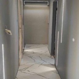 NEW DESIGN KAMIL SZAREK - Korytarz w trakcie wykańczania: położone marmurowe płytki podłogowe, szare ściany, oświetlenie LED wzdłuż ścian i sufitu. Widoczne narzędzia glazurnicze.