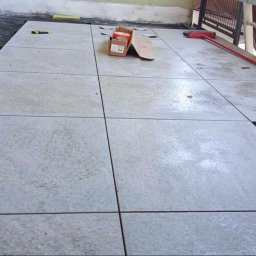 NEW DESIGN KAMIL SZAREK - Jasne, kwadratowe płytki ułożone na balkonie, widoczne narzędzia i materiały budowlane. Częściowo ukończona praca, z czarną folią pod płytkami.