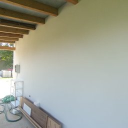 NEW DESIGN KAMIL SZAREK - Ściana po malowaniu z drewnianą konstrukcją sufitu. Na podłodze leżą płyty i narzędzia, sugerujące prace wykończeniowe.