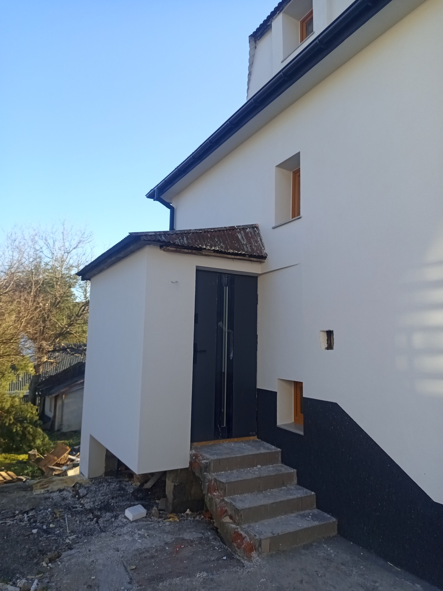 Odnowiona elewacja budynku z nowym wejściem i schodami. Świeże, białe tynki kontrastują z ciemnym wykończeniem dolnej części ściany. Widoczne detale okien i dachu.