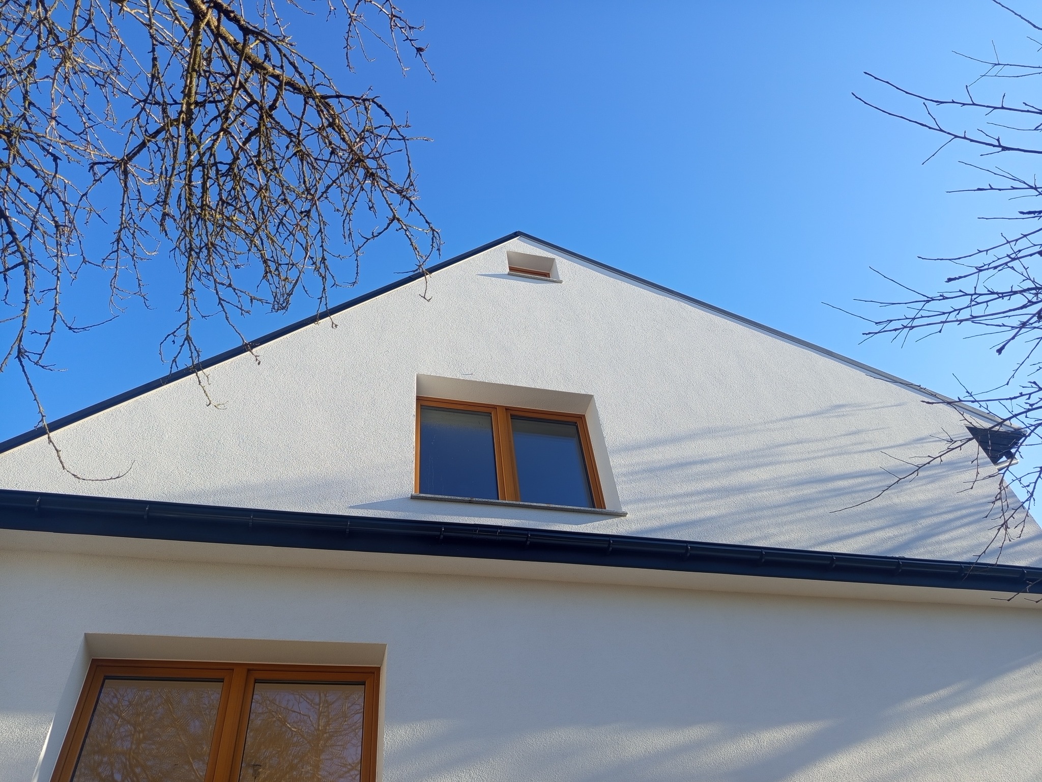 Biała elewacja domu z drewnianymi oknami i czarną rynną na tle błękitnego nieba, z gałęziami drzew w rogu kadru. Widok z dołu.