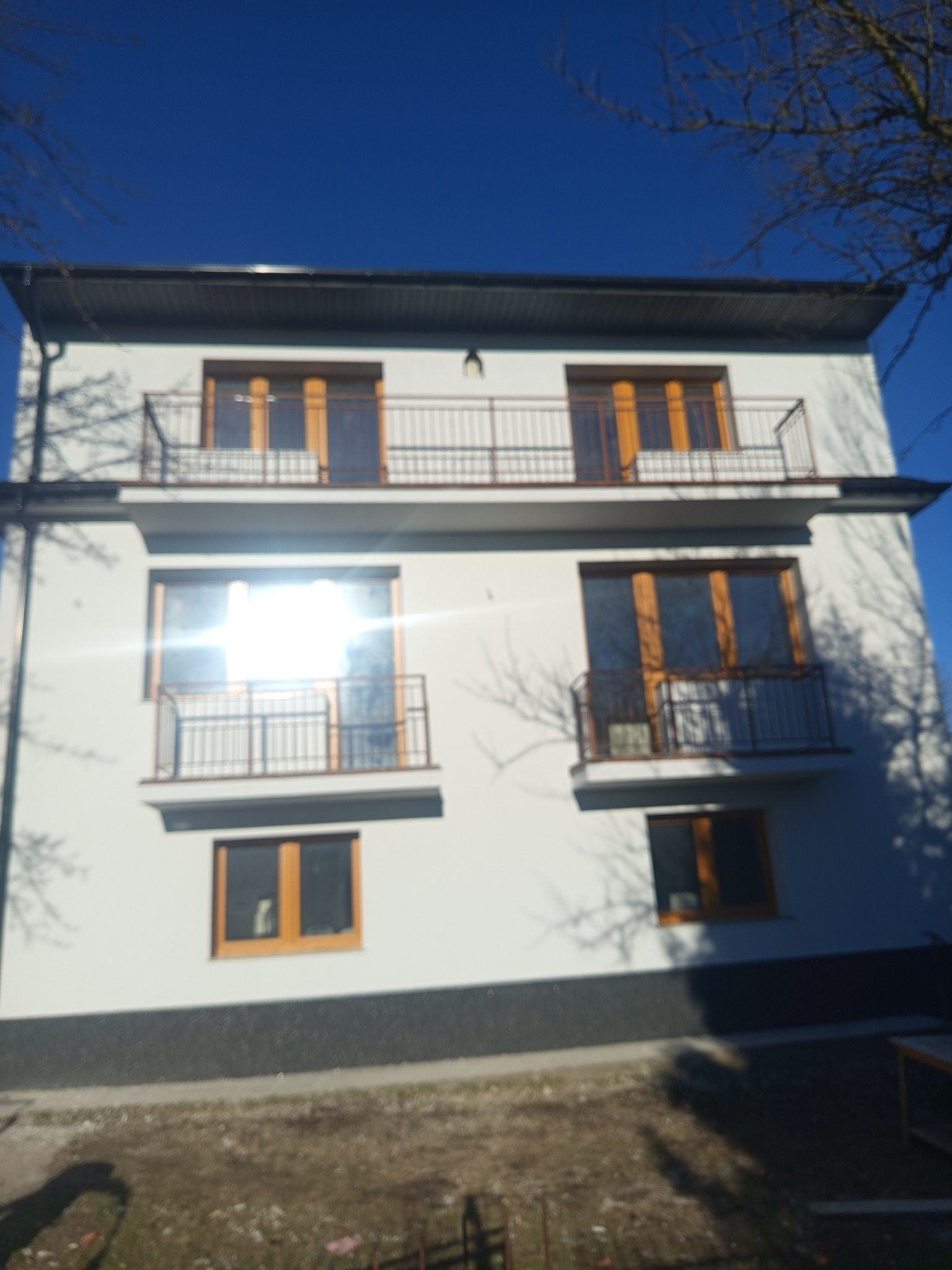 Biały, dwupiętrowy dom z drewnianymi oknami i balkonami z czarną balustradą, widziany z dołu w słoneczny dzień. Widoczne nagie gałęzie drzew.