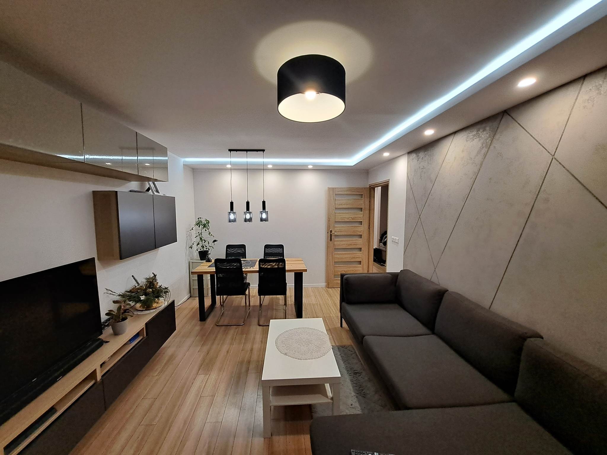 Nowoczesny salon z szarą sofą narożną, drewnianą podłogą i geometryczną ścianą. Centralnie umieszczony stół jadalny z czarnymi krzesłami. Oświetlenie sufitowe LED.