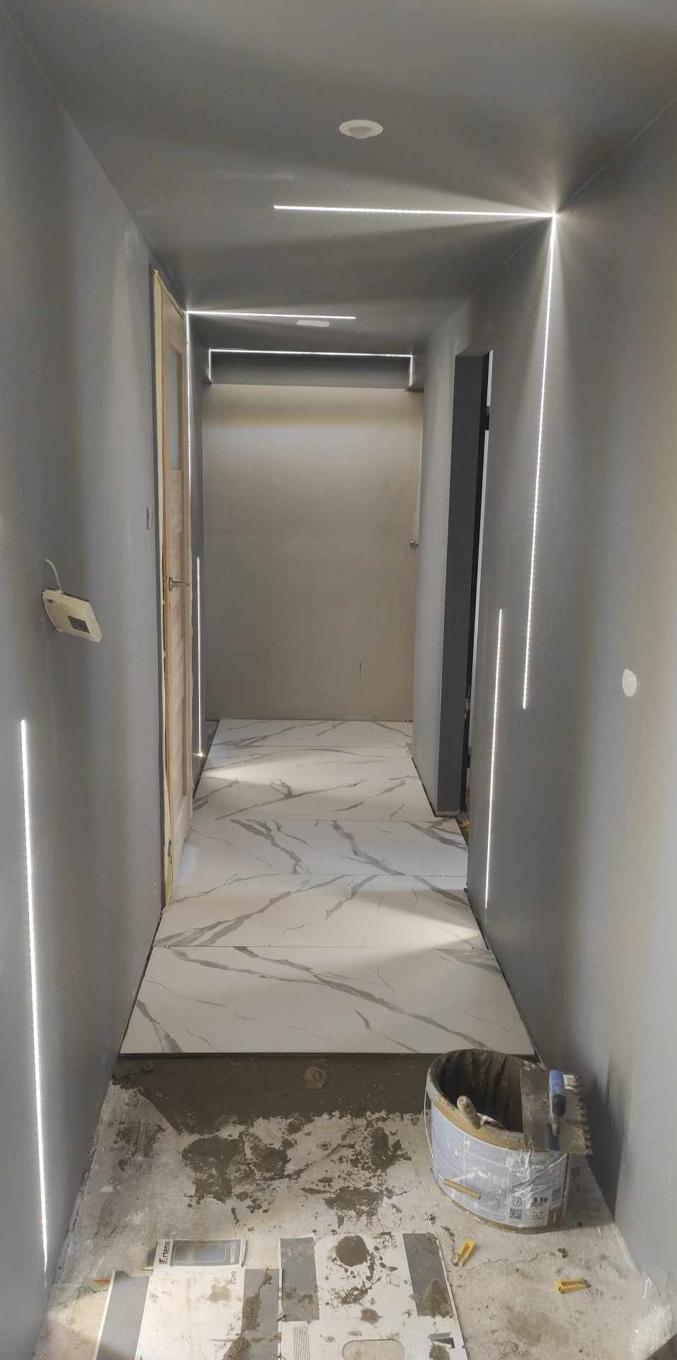 Korytarz w trakcie wykańczania: położone marmurowe płytki podłogowe, szare ściany, oświetlenie LED wzdłuż ścian i sufitu. Widoczne narzędzia glazurnicze.