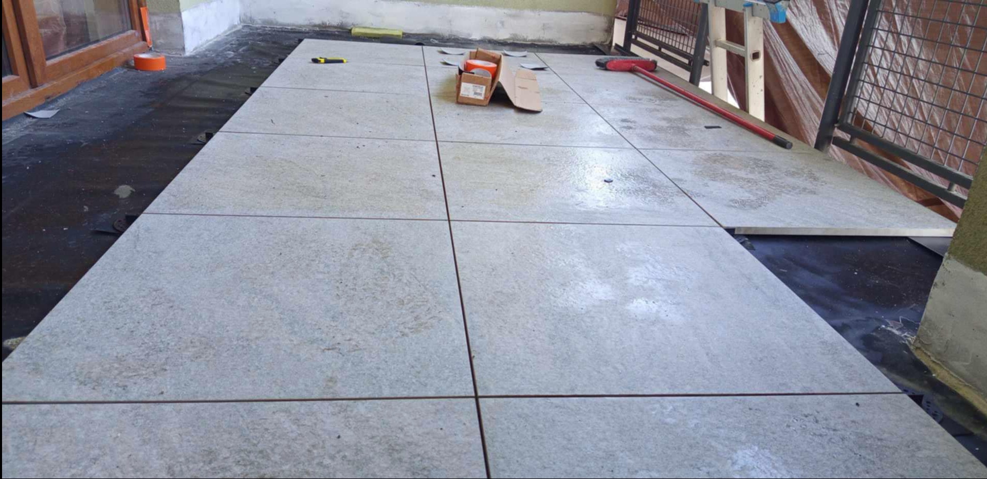 Jasne, kwadratowe płytki ułożone na balkonie, widoczne narzędzia i materiały budowlane. Częściowo ukończona praca, z czarną folią pod płytkami.