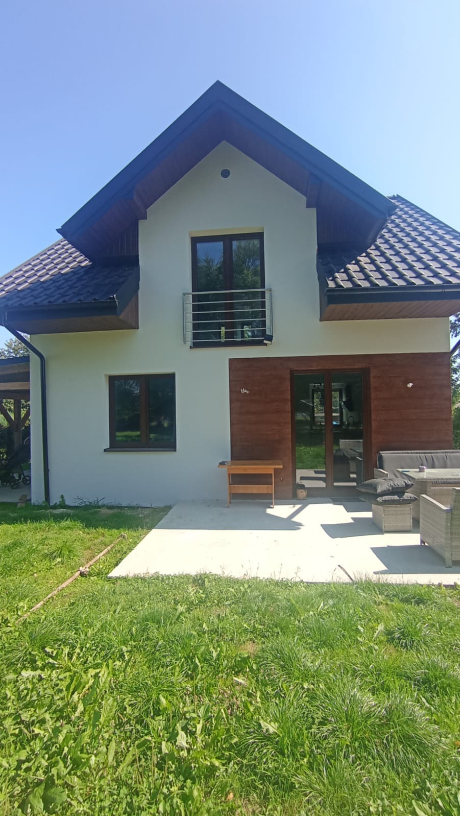 Dwukondygnacyjny dom z balkonem i drewnianą elewacją, widok z trawnika w słoneczny dzień. Widoczne meble ogrodowe i fragment tarasu.