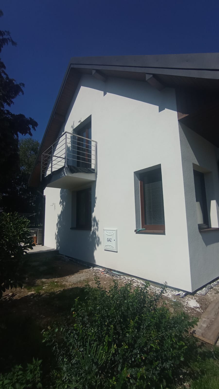 Biała elewacja domu jednorodzinnego z balkonem i oknami w brązowych ramach, widok z poziomu ogrodu w słoneczny dzień. Skrzynka gazowa na ścianie.