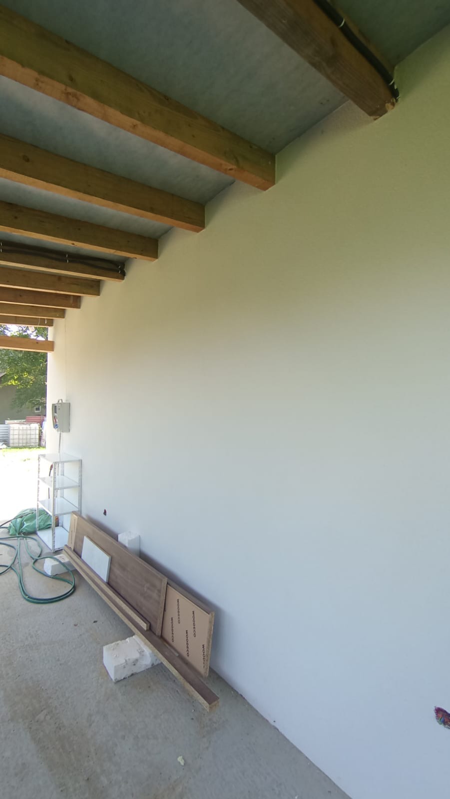 Ściana po malowaniu z drewnianą konstrukcją sufitu. Na podłodze leżą płyty i narzędzia, sugerujące prace wykończeniowe.