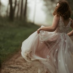 Unsimple Photography - Kobieta w koronkowej sukni ślubnej z pudrowo-różowym tiulem, biegnąca leśną ścieżką w tenisówkach, unosząc brudny dół sukni.