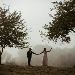 Unsimple Photography - Para młoda trzymająca się za ręce, pozująca na wzgórzu w mglisty dzień, otoczona drzewami.