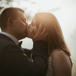 Unsimple Photography - Młoda para w pocałunku podczas sesji plenerowej, on w granatowym garniturze z muchą, ona w koronkowej sukni ślubnej, delikatne światło słoneczne w tle.