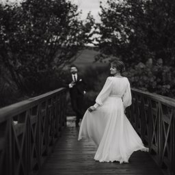 Unsimple Photography - Czarno-białe zdjęcie panny młodej w zwiewnej sukni, odwracającej się na drewnianym moście, z panem młodym w tle, otoczone drzewami.