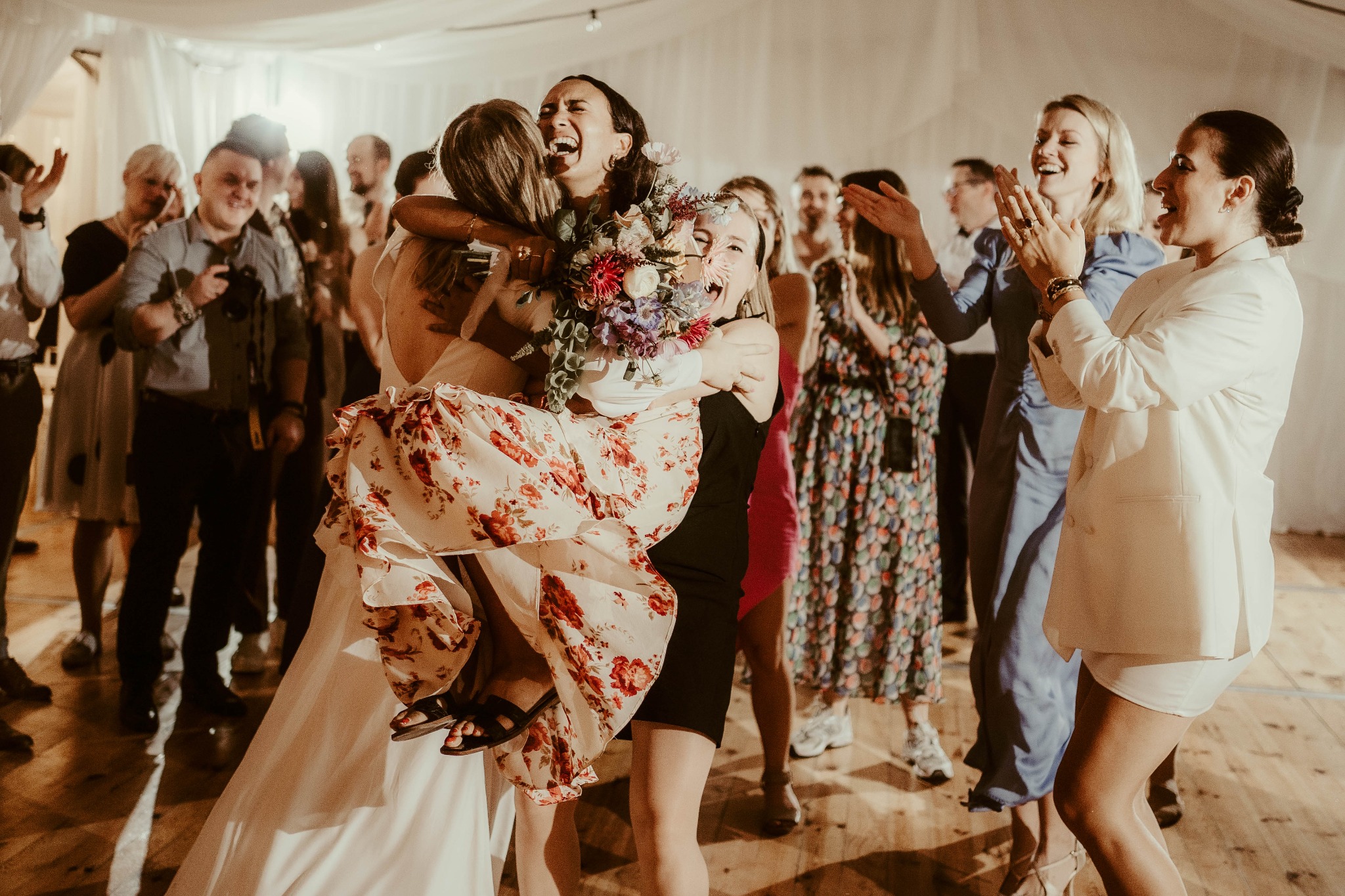 Panna młoda w białej sukni niesiona na rękach przez kobietę w czarnej sukience, śmiech i radość, bukiet kwiatów w dłoniach panny młodej, goście weselni klaszczą w tle, sala weselna z drewnianą...