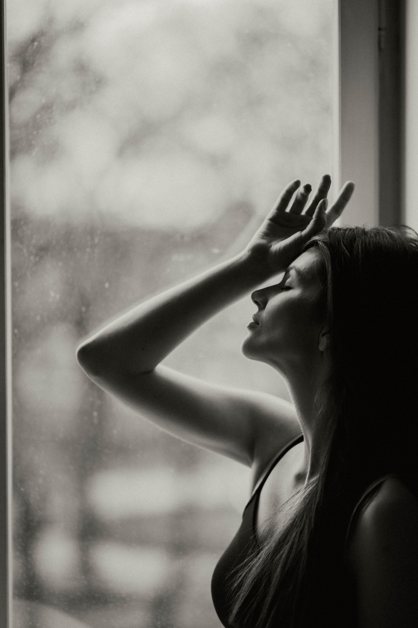 Monochromatyczny portret kobiety opierającej dłoń o czoło, profil twarzy i długie włosy na tle okna z kroplami deszczu.